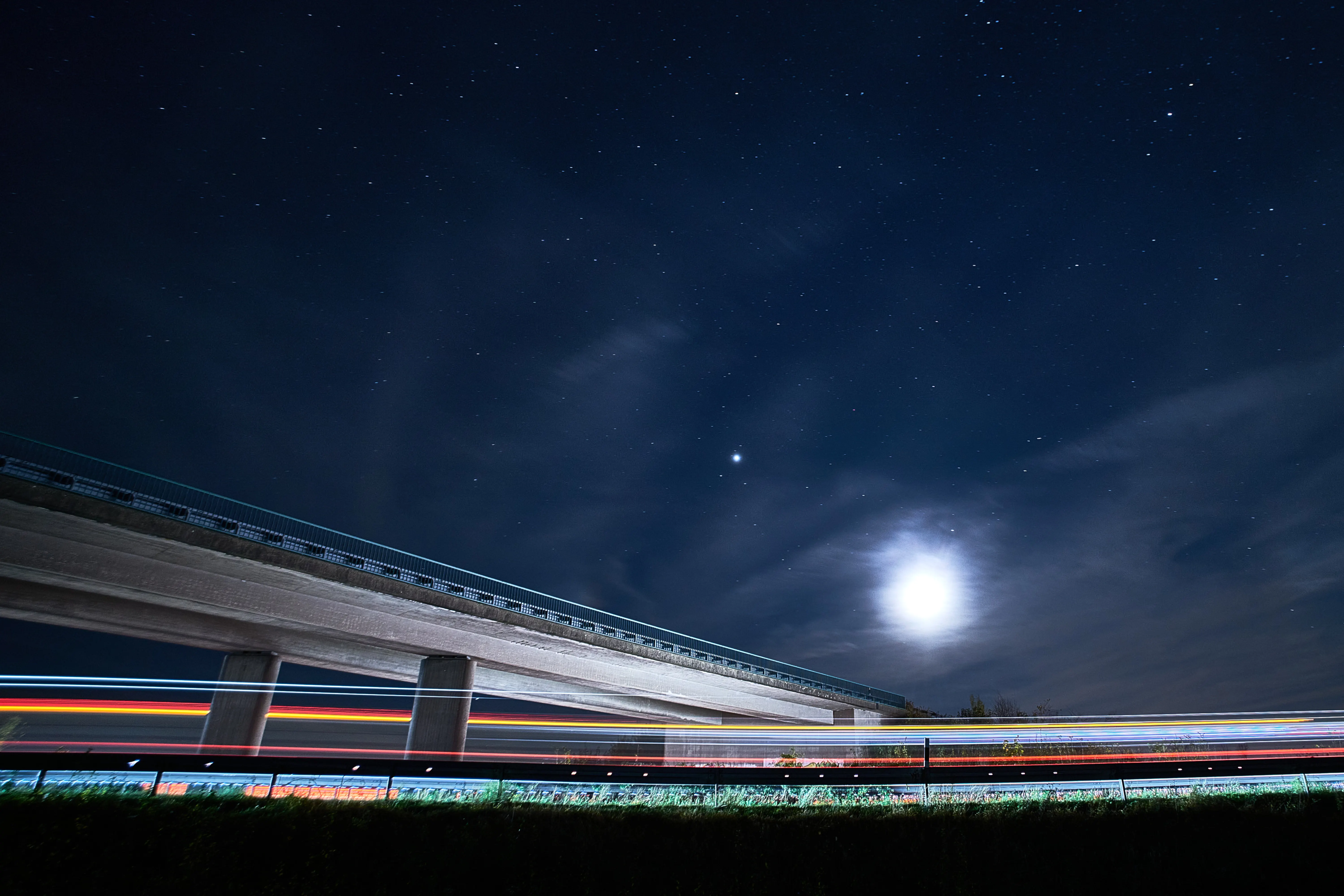 Autobahnbrücke bei Nacht unter Sternenhimmel, bunte Lichtspuren vorbeifahrender Autos im Vollmondlicht