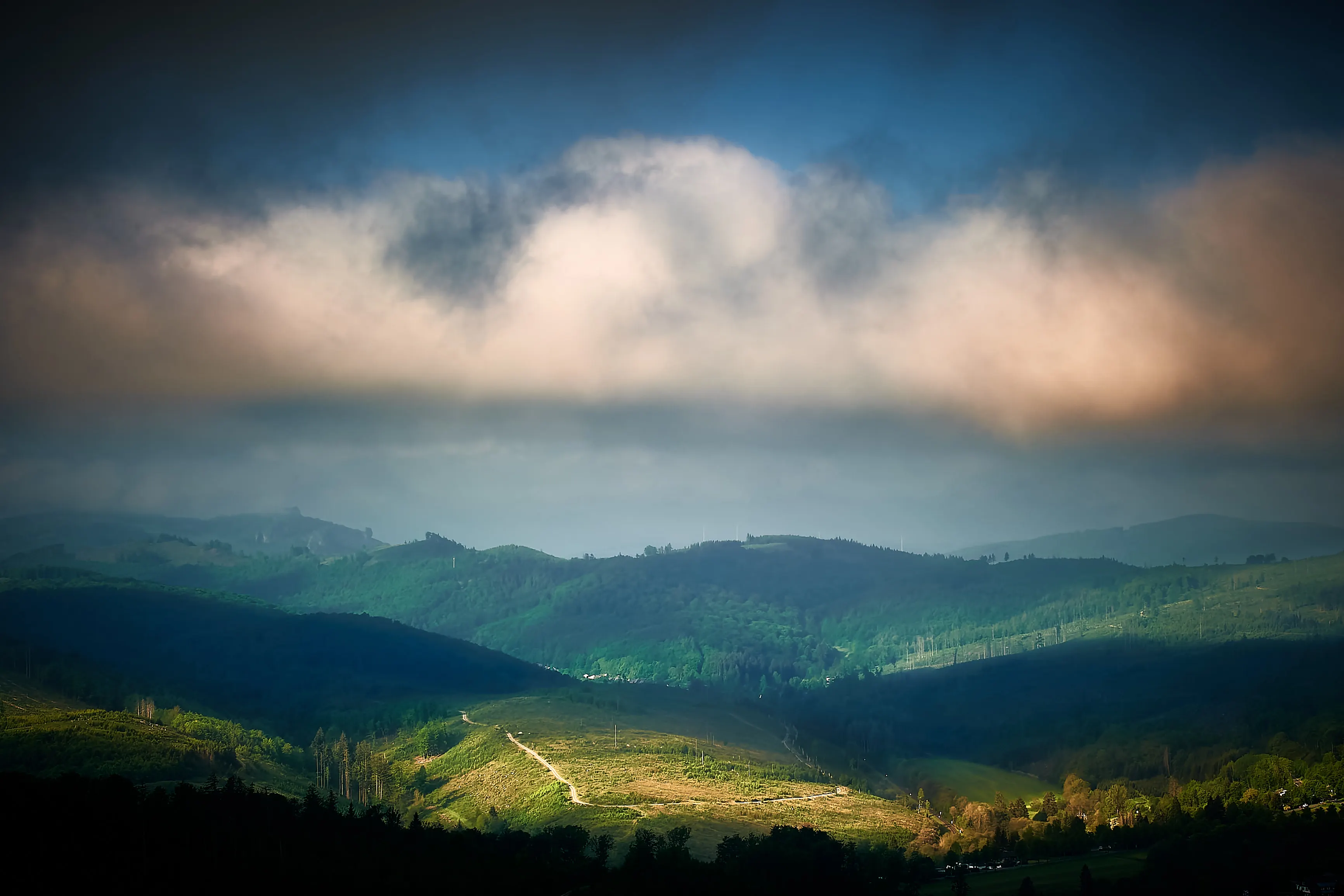 Tief hängende Wolken über bewaldeten Hügeln, eine sonnenbeschienene grüne Lichtung im Tal darunter