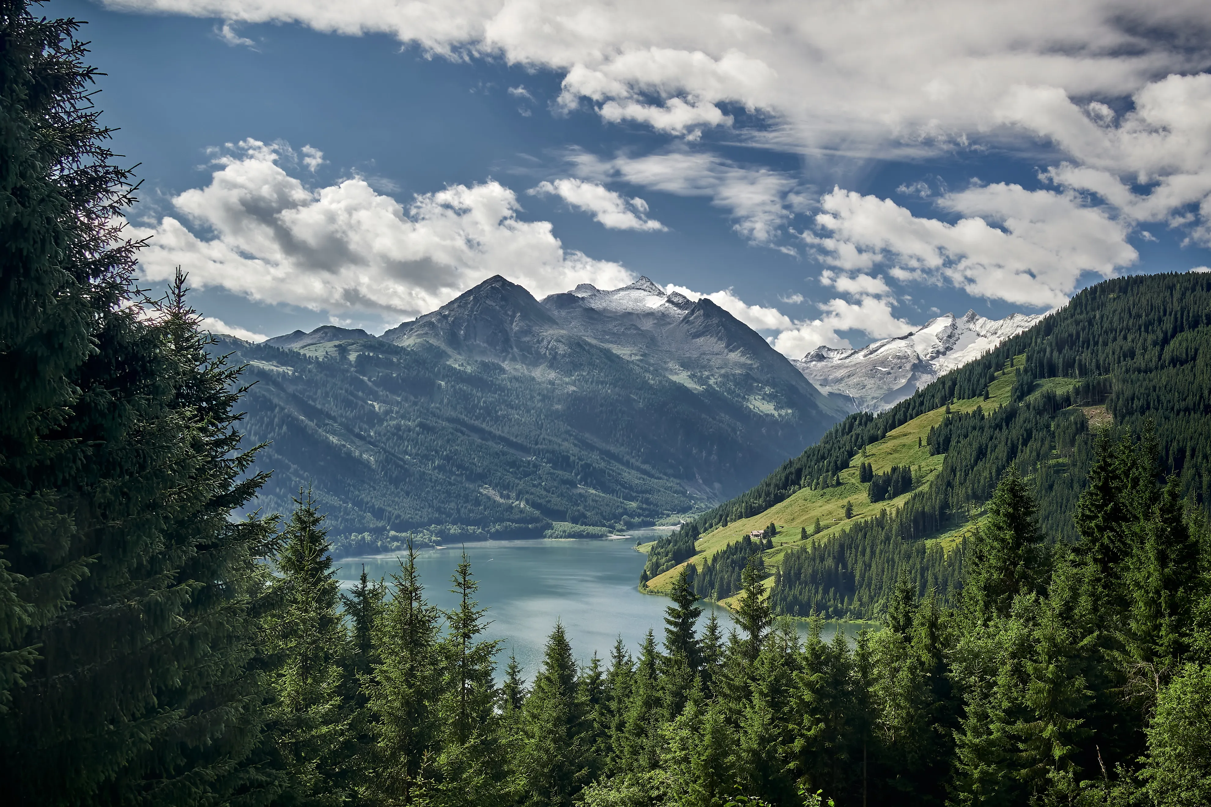 Türkisfarbener Bergsee zwischen bewaldeten Hängen, dahinter schneebedeckte und vergletscherte Gipfel