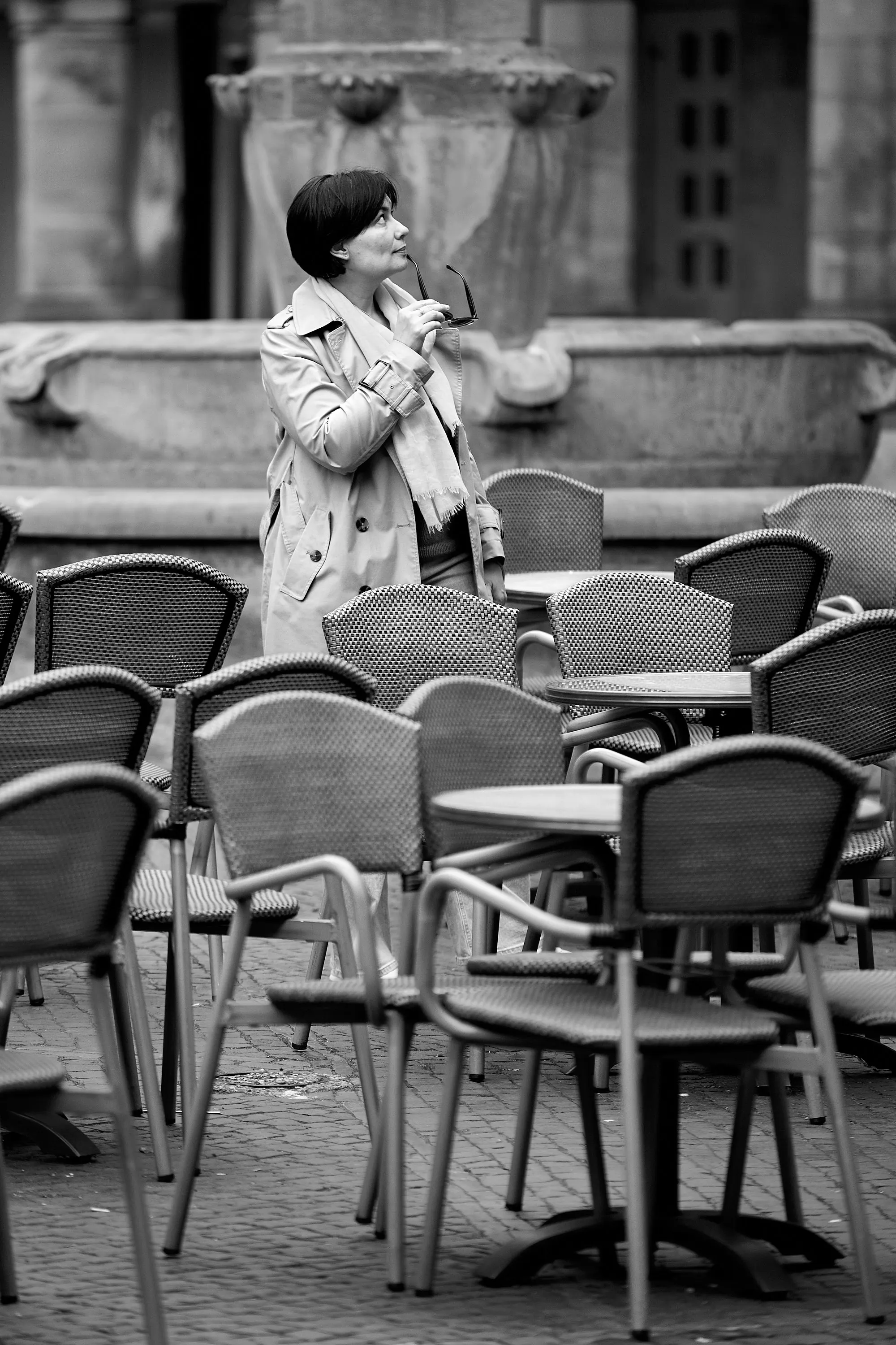Schwarzweiß-Aufnahme einer Frau im Trenchcoat zwischen den leeren Stühlen eines Straßencafés