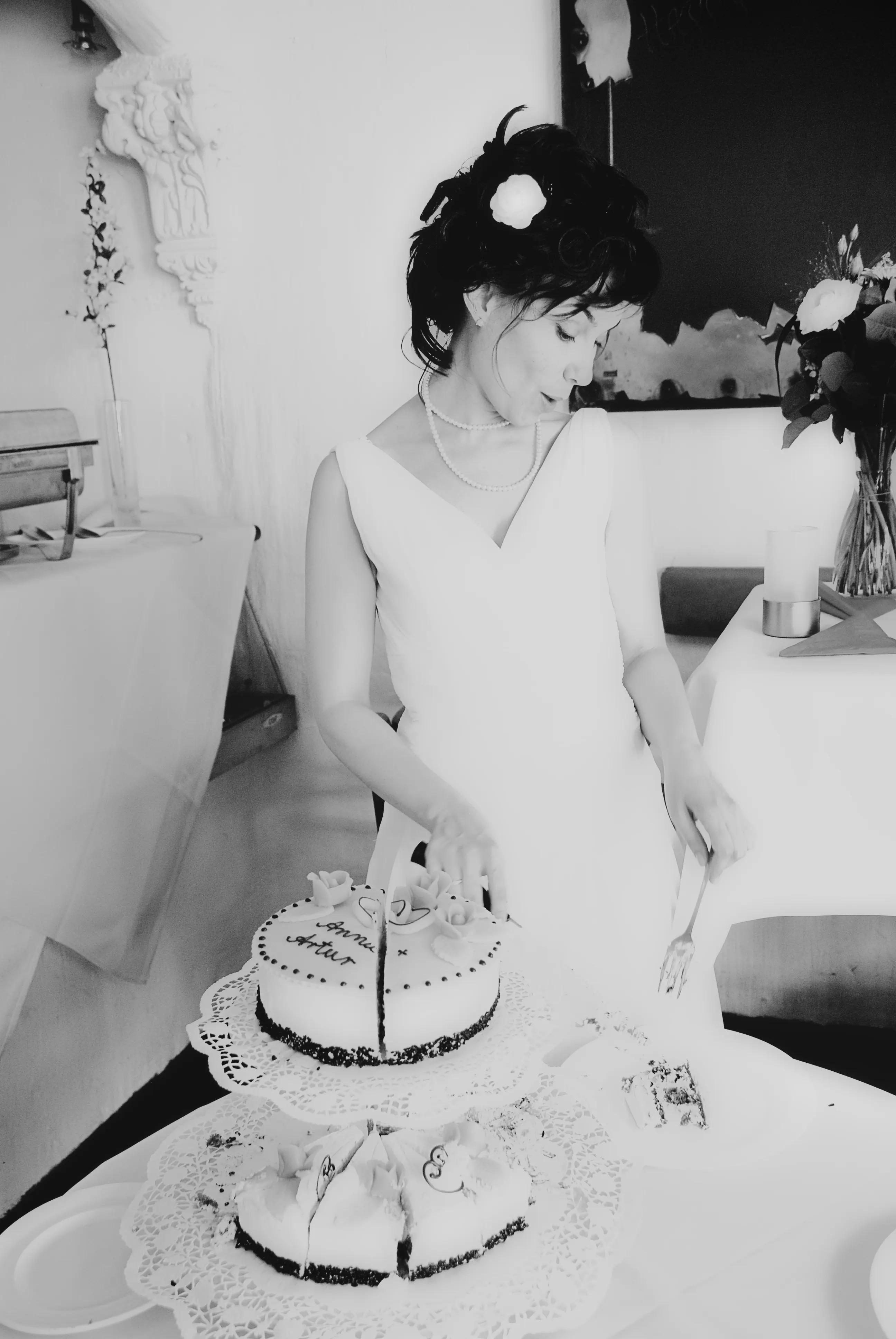 Schwarzweiß-Aufnahme einer Braut mit Blüte im Haar, die eine herzförmige Hochzeitstorte auf der Etagere anschneidet