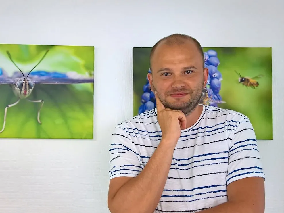 Porträt von Artur, der lächelnd das Kinn auf die Hand stützt, vor Naturfotografien an der Wand