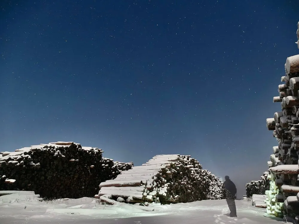 Verschneite Holzstapel unter dem Sternenhimmel, eine einzelne Person steht im Schnee davor